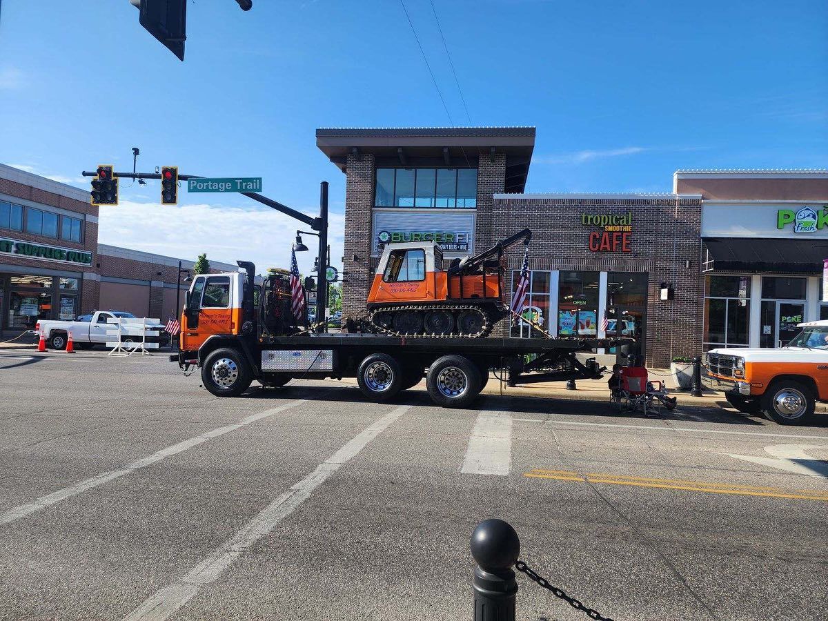 Orange flatbed truck carrying an orange excavator on a city street. Buildings and a white truck are nearby.
