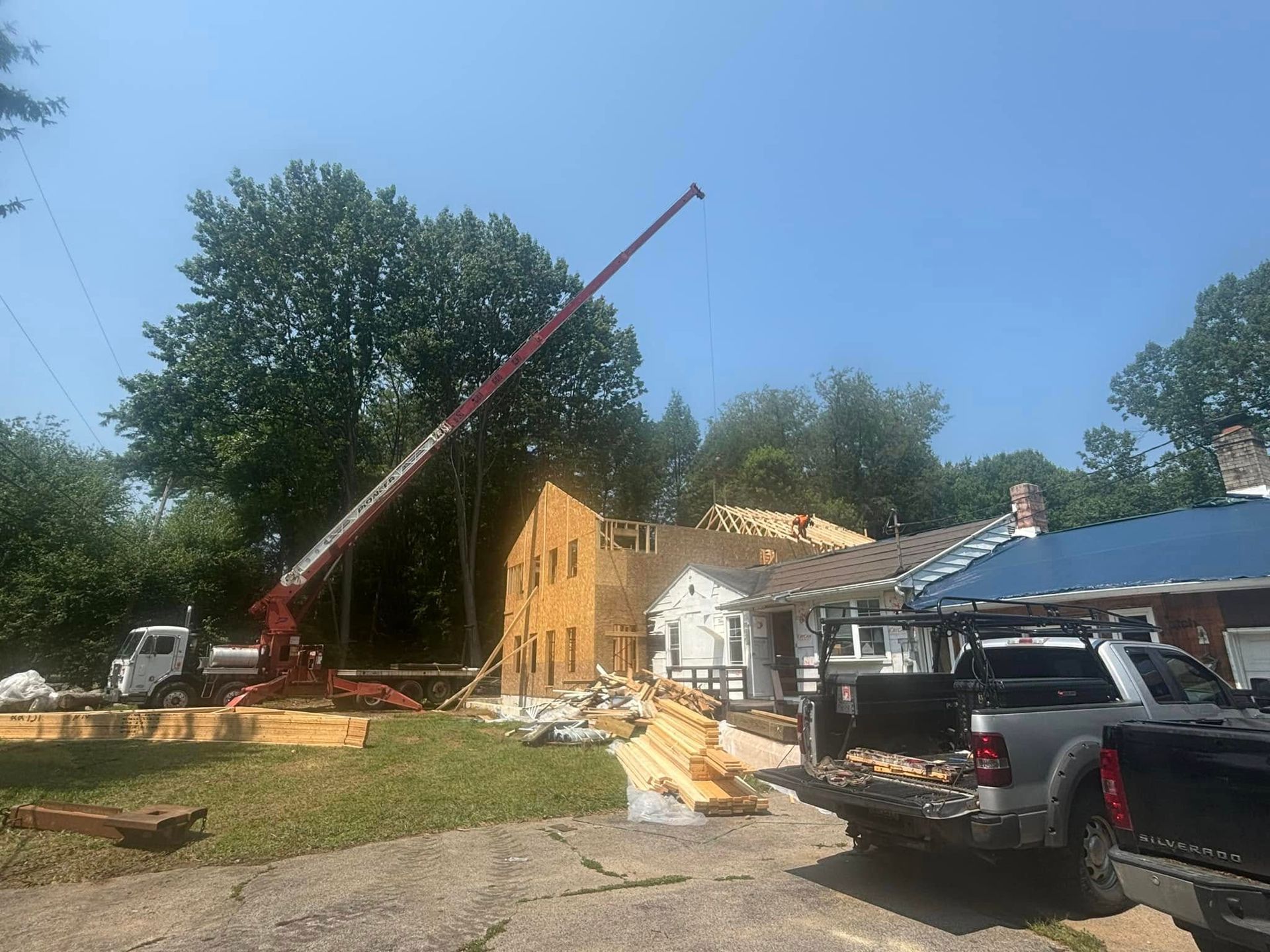 Construction site with crane lifting materials to a house. Trucks, lumber, and trees are also visible under a blue sky.