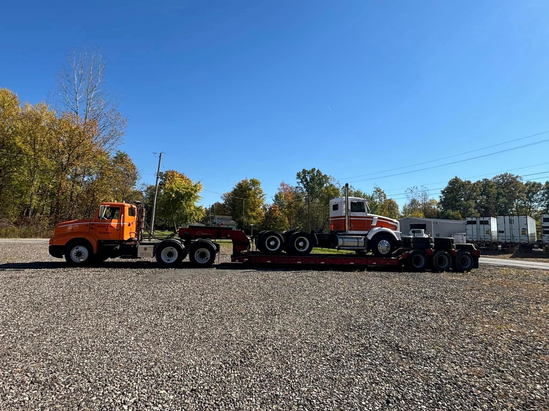 Orange and white semi-trucks on a lowboy trailer, parked on gravel. Blue sky, trees in the background.