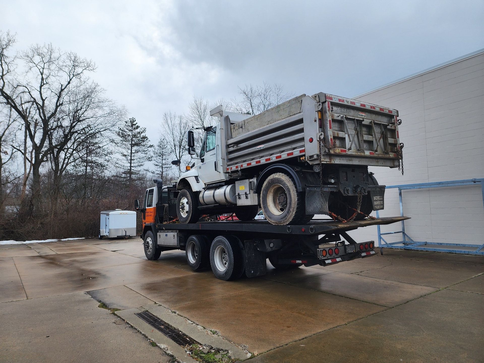 Flatbed tow truck carrying a white dump truck, on a wet concrete surface under an overcast sky.