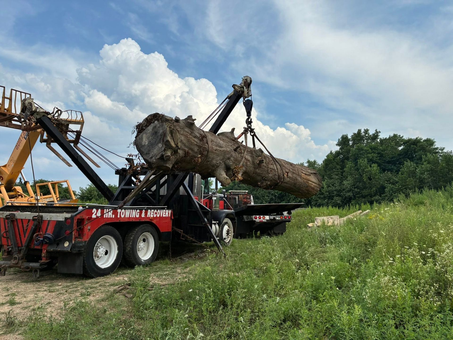 A large tree trunk being lifted by a red tow truck on a sunny day in a grassy field.