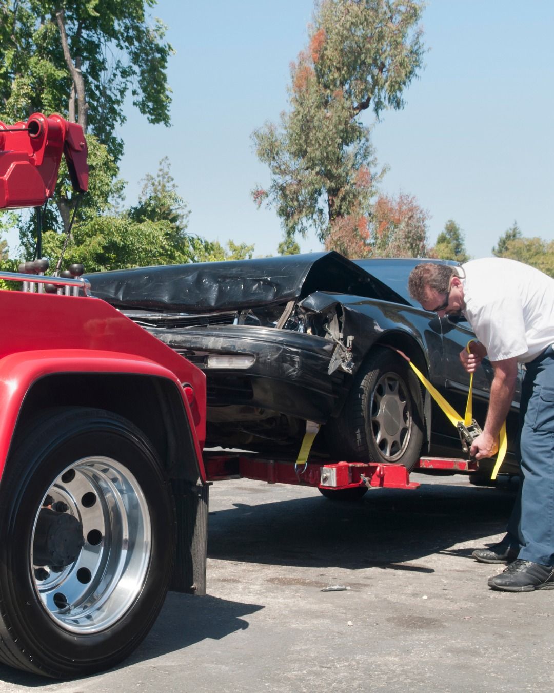 Tow truck loading a heavily damaged black car. A man secures straps. Sunny outdoor scene.