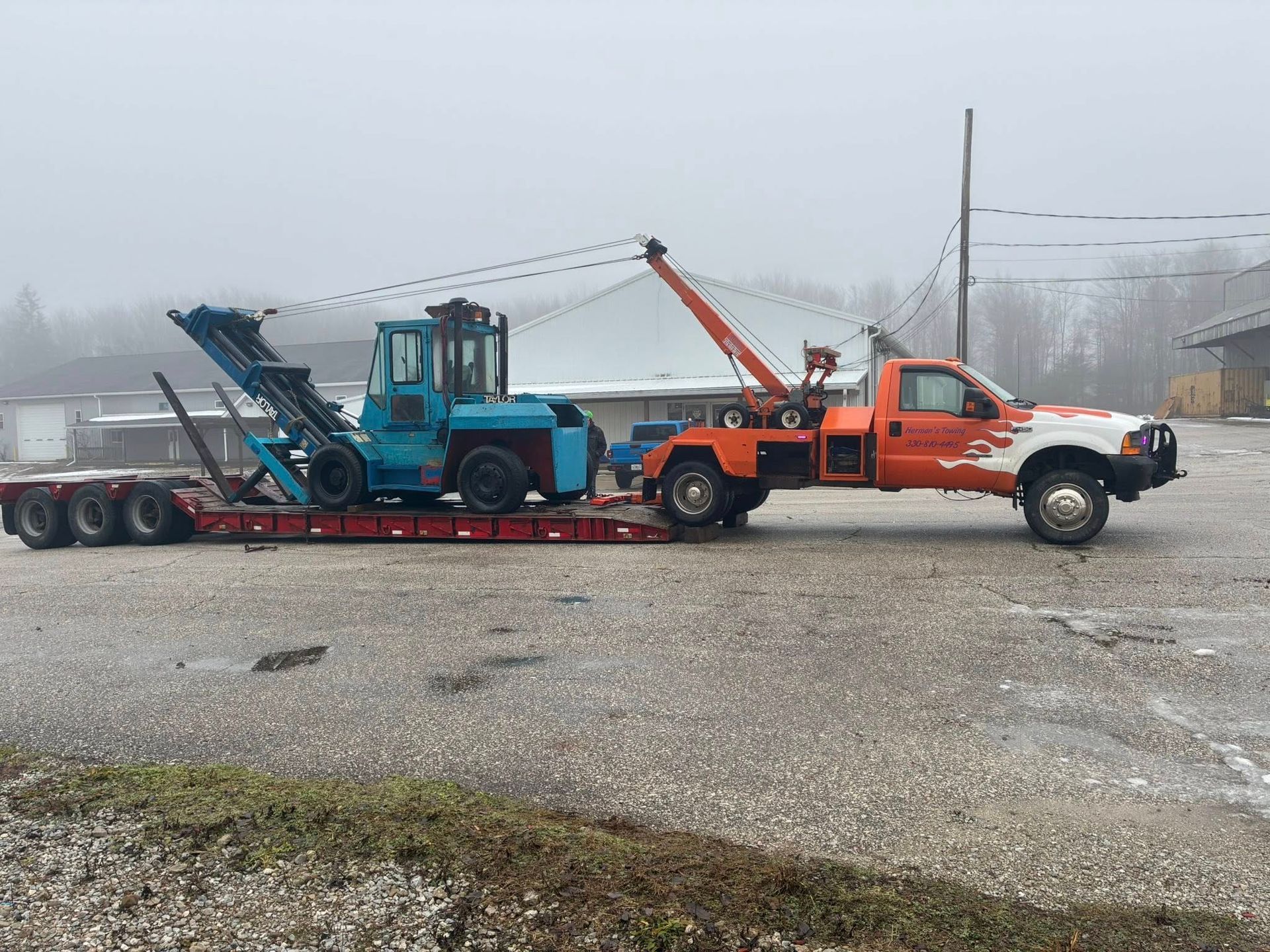 Orange tow truck hauling a blue forklift on a flatbed trailer in a misty outdoor setting.