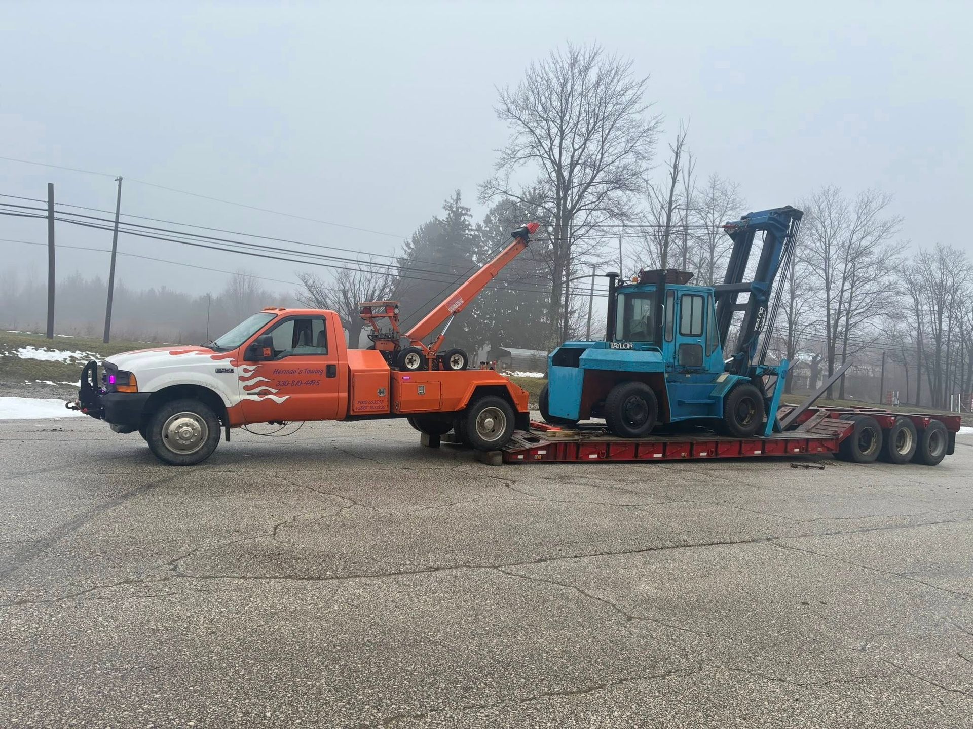 Orange tow truck hauling a blue forklift on a flatbed trailer on a gray road.