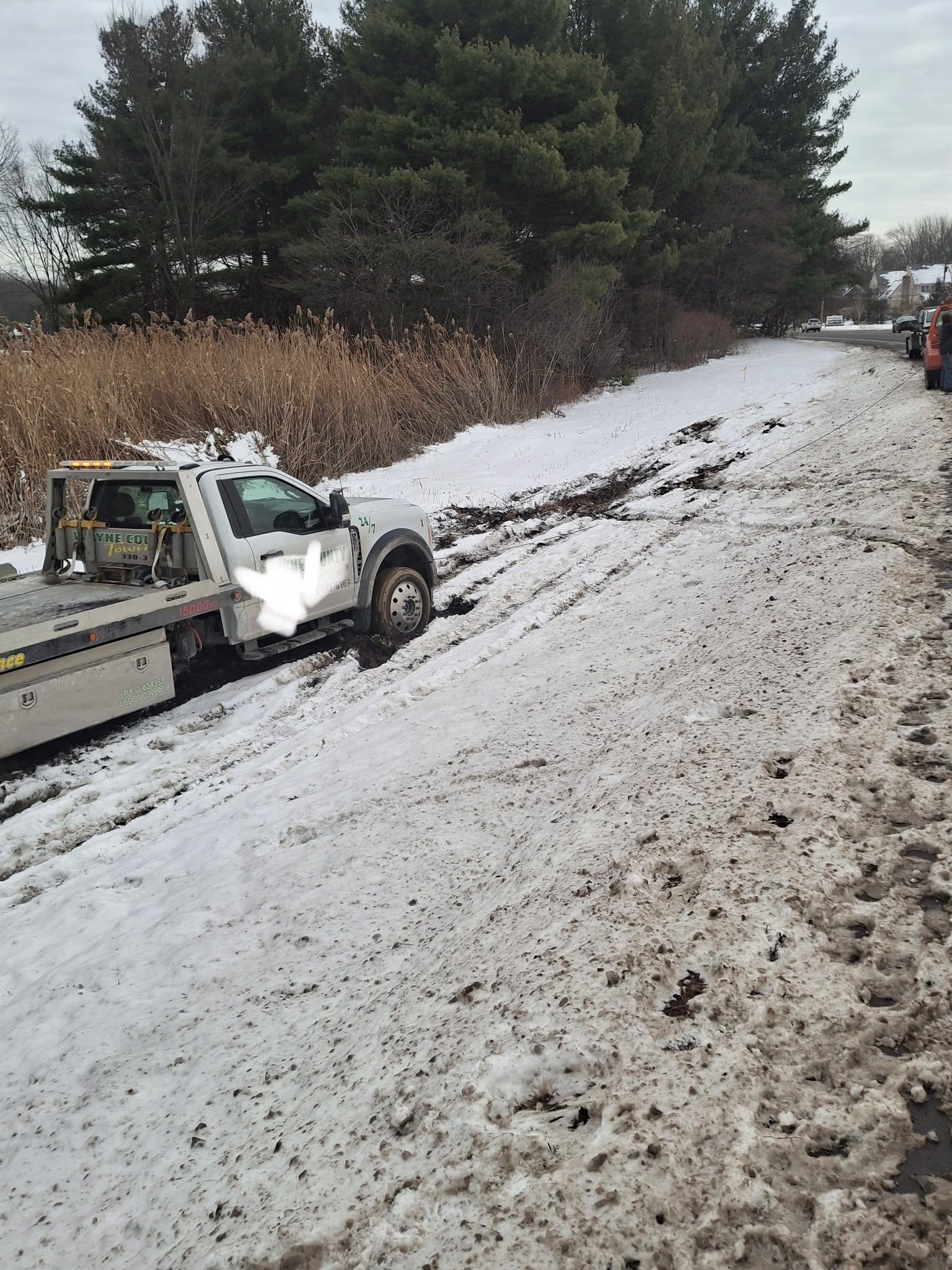 Tow truck partially stuck in snow-covered mud beside a road, trees in the background.