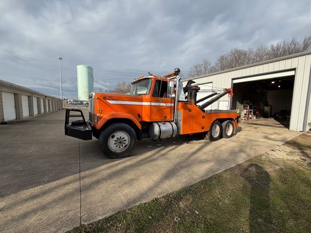 An orange tow truck parked on a concrete lot in front of a long, white metal storage building under a cloudy sky.