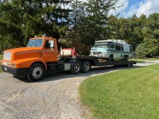 An orange semi-truck transports a vintage white and blue camper van on a flatbed trailer on a gravel driveway.