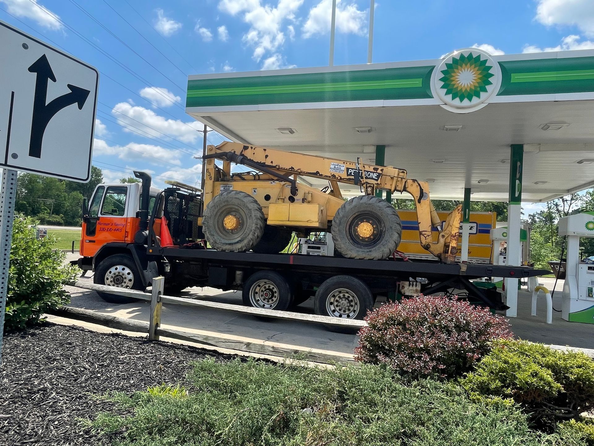 A flatbed truck carrying a large yellow construction machine at a BP gas station.
