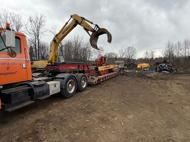 An orange semi-truck transports a yellow excavator with a claw attachment on a flatbed trailer in a dirt lot.