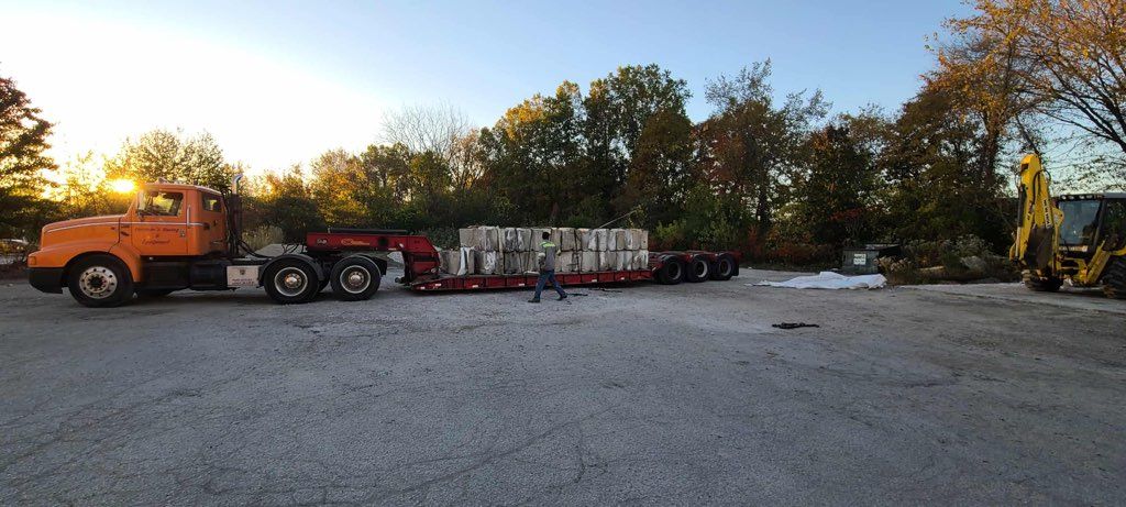 An orange truck hitched to a flatbed trailer loaded with construction materials, parked on a gravel lot at sunset.