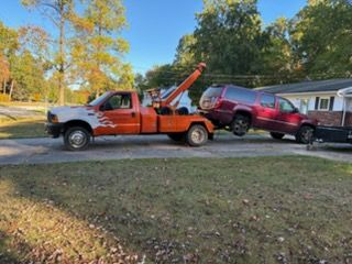Truck loading large log on a trailer in a grassy area with trees and blue sky.