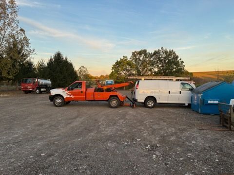 An orange tow truck is towing a white cargo van in a gravel lot, with a blue dumpster and another truck nearby at dusk.