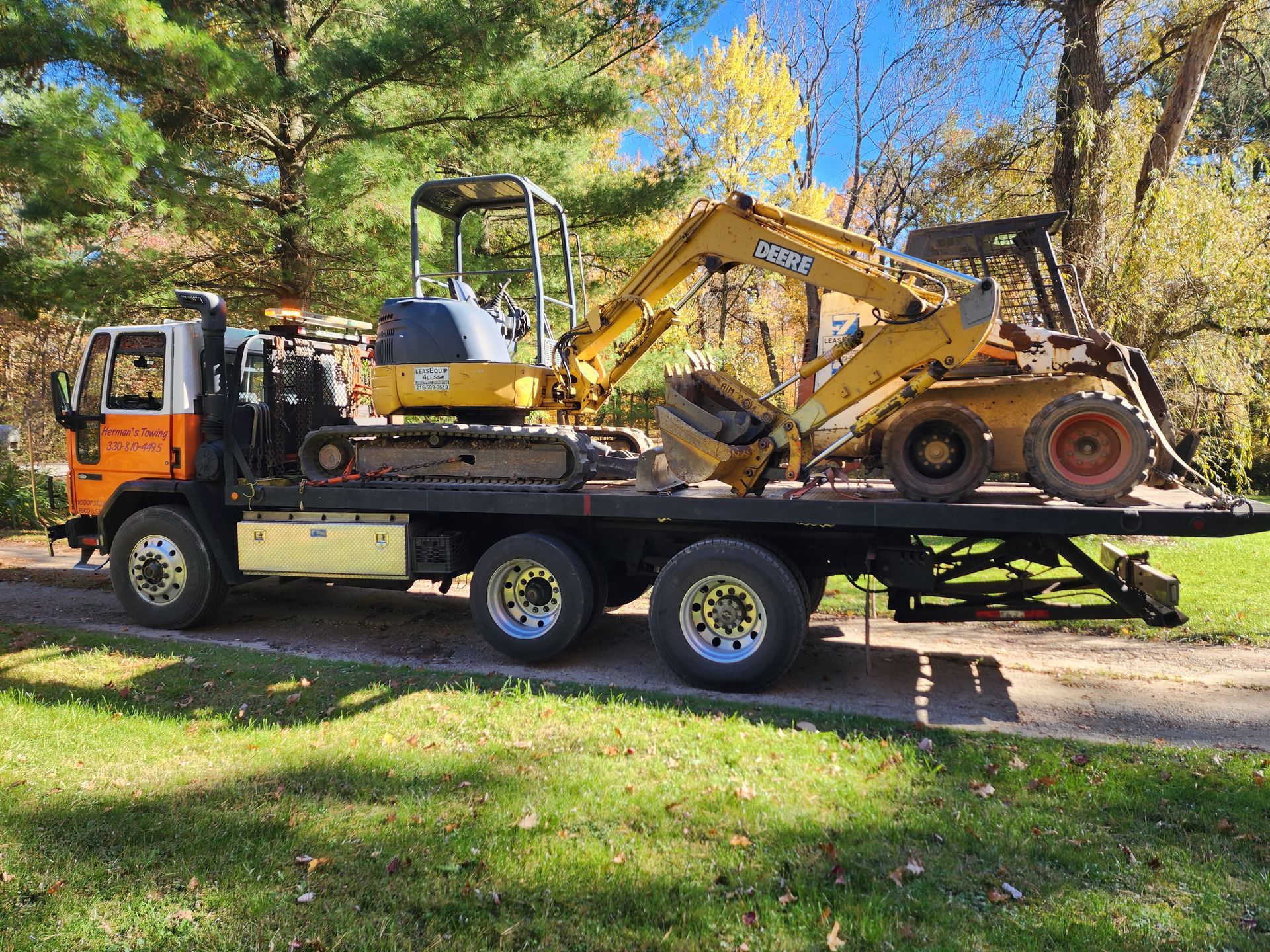 Flatbed truck carrying a yellow excavator and a front-end loader on a grassy area, trees in the background.