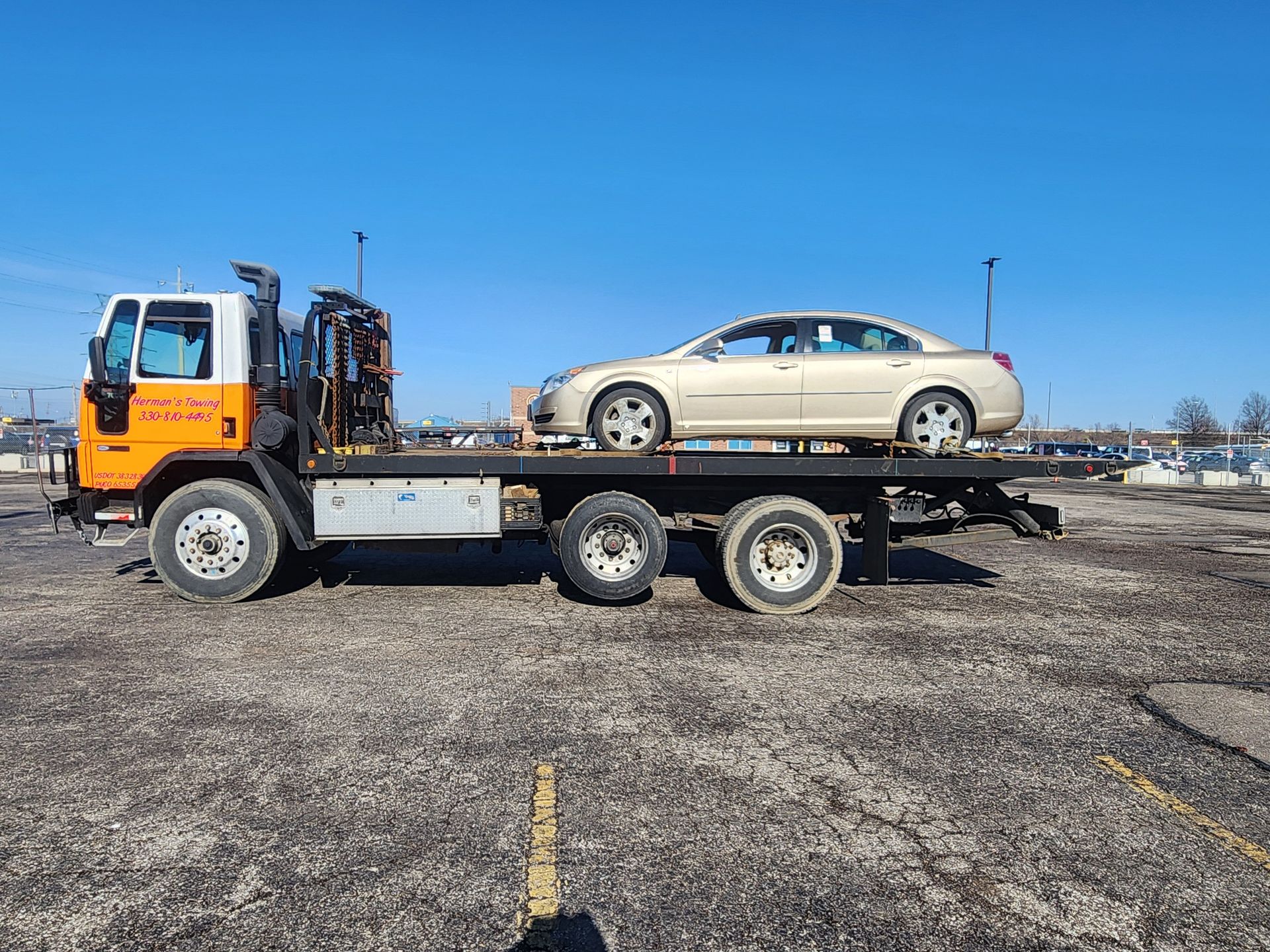 Yellow tow truck carrying a white camper van on a gravel lot under a cloudy sky.