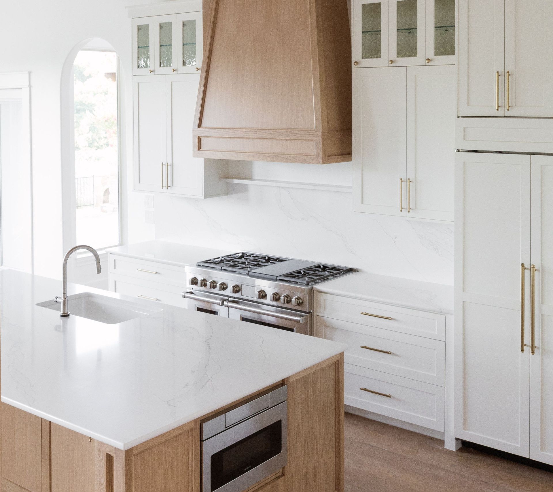 Modern white kitchen with light wood accents, island, stainless steel appliances, and a large range hood.