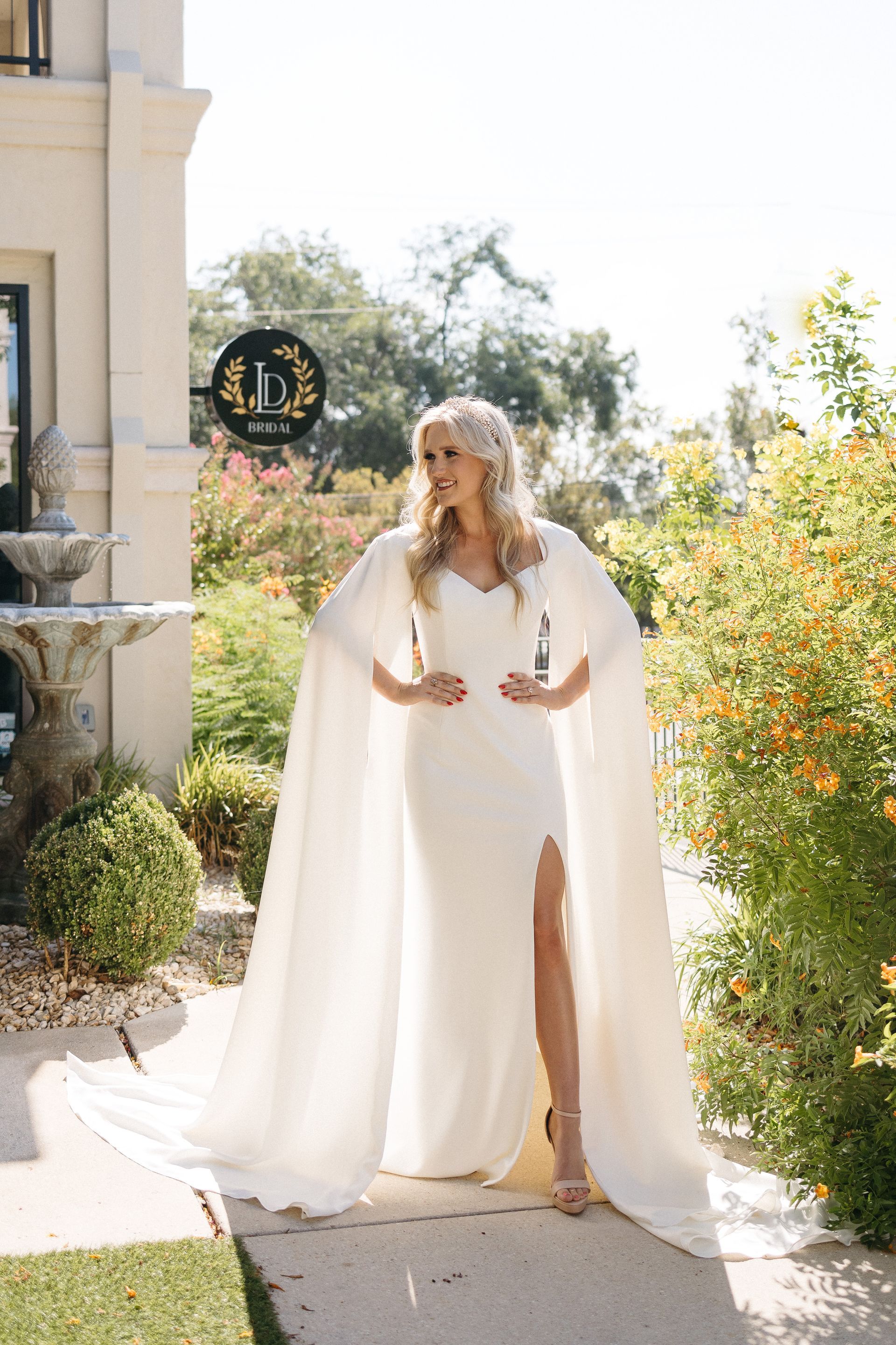 A woman in a white wedding dress is standing on a sidewalk holding two bottles of wine.
