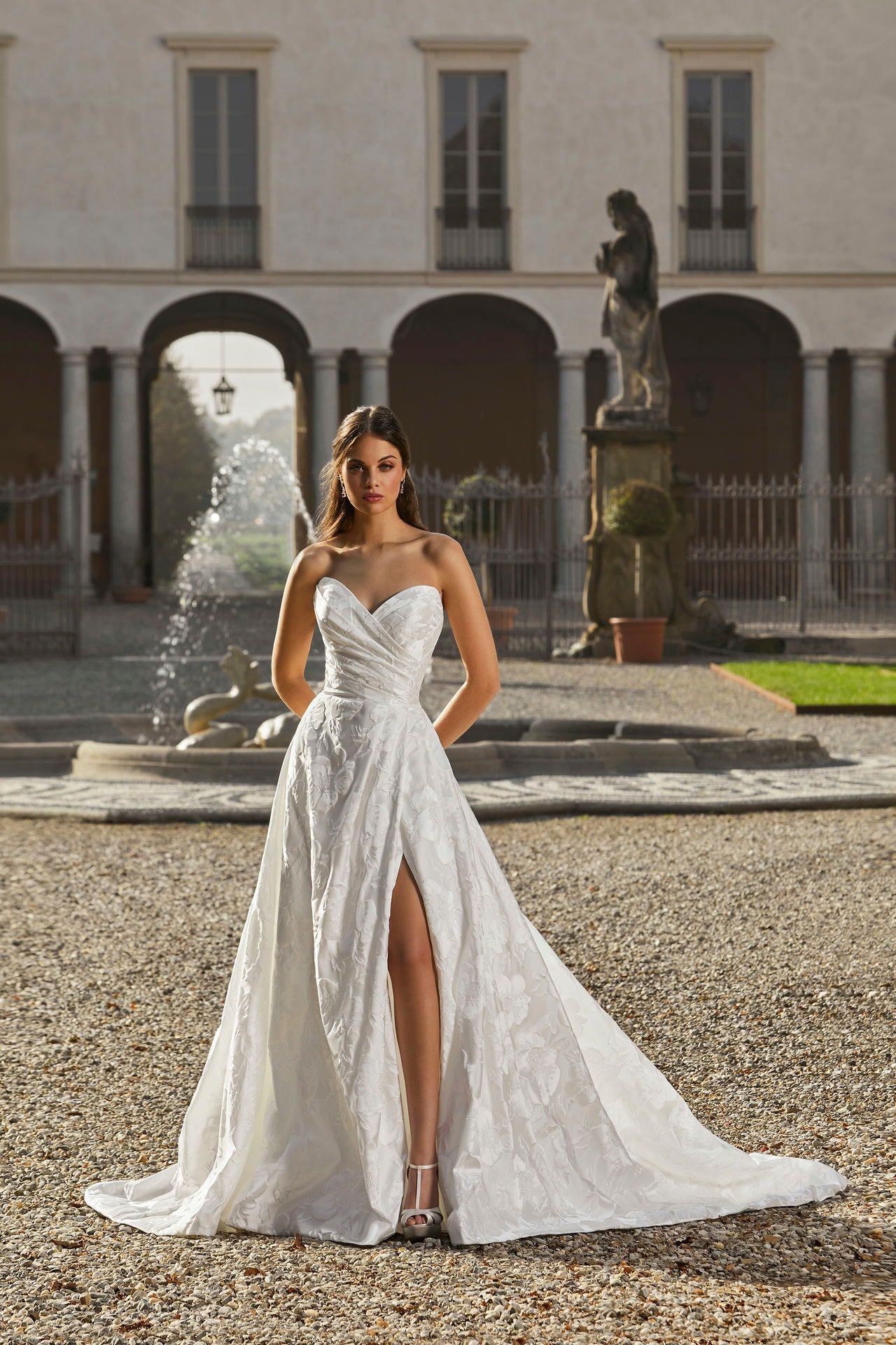 A woman in a white wedding dress is standing in front of a fountain.