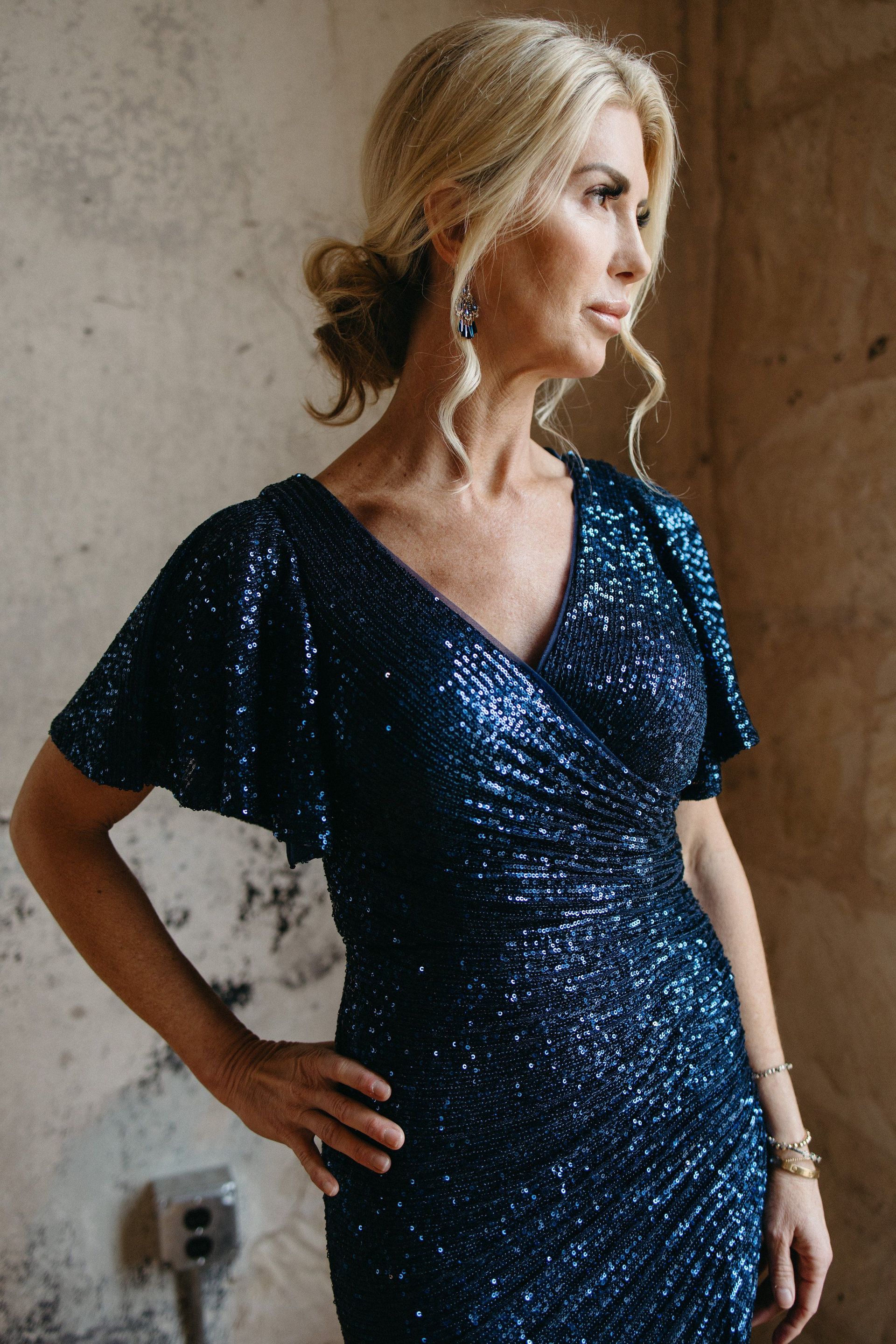 A woman in a blue sequined dress is standing in front of a wall.