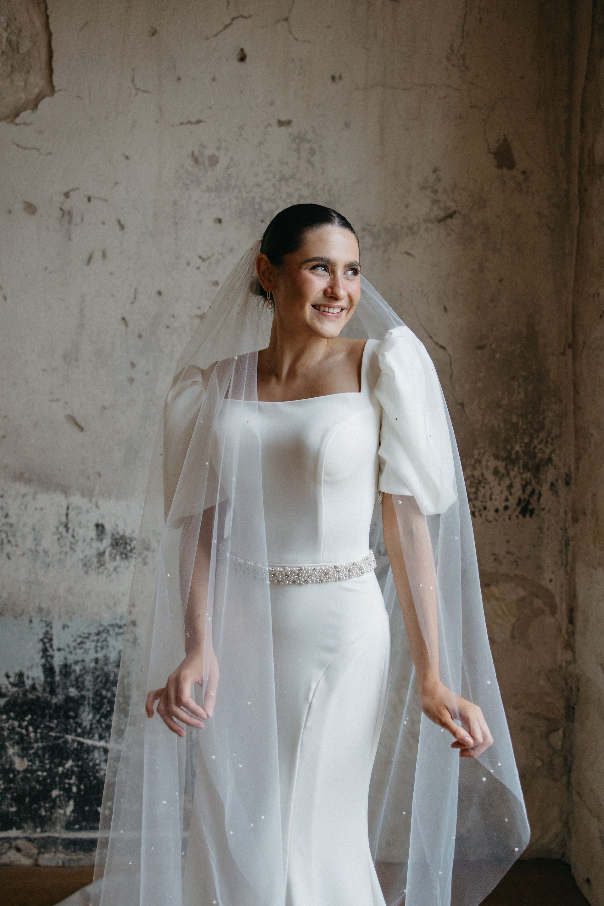 A woman in a white dress and veil is standing in front of a wall.