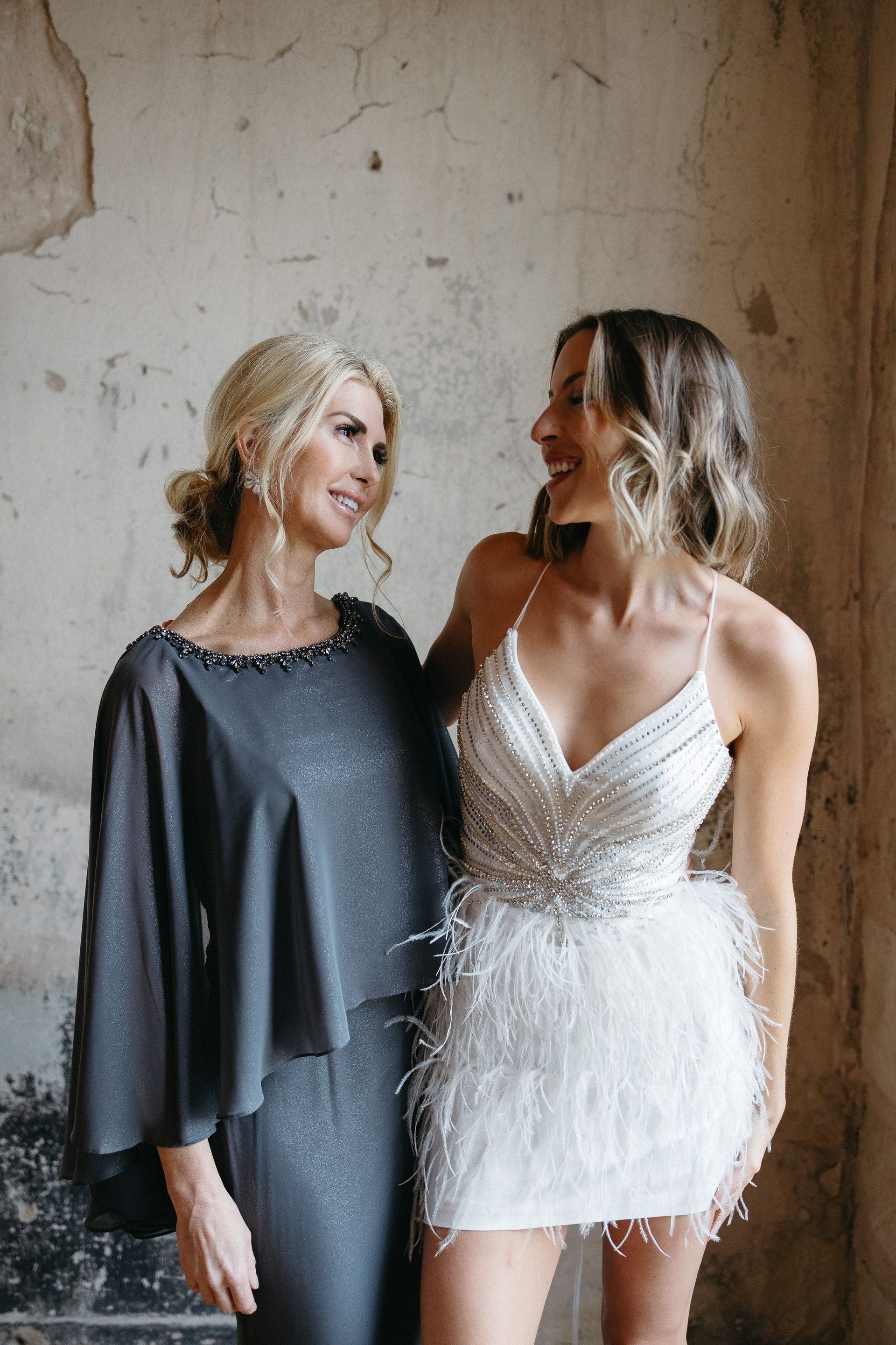 Two women in dresses are standing next to each other in front of a wall.