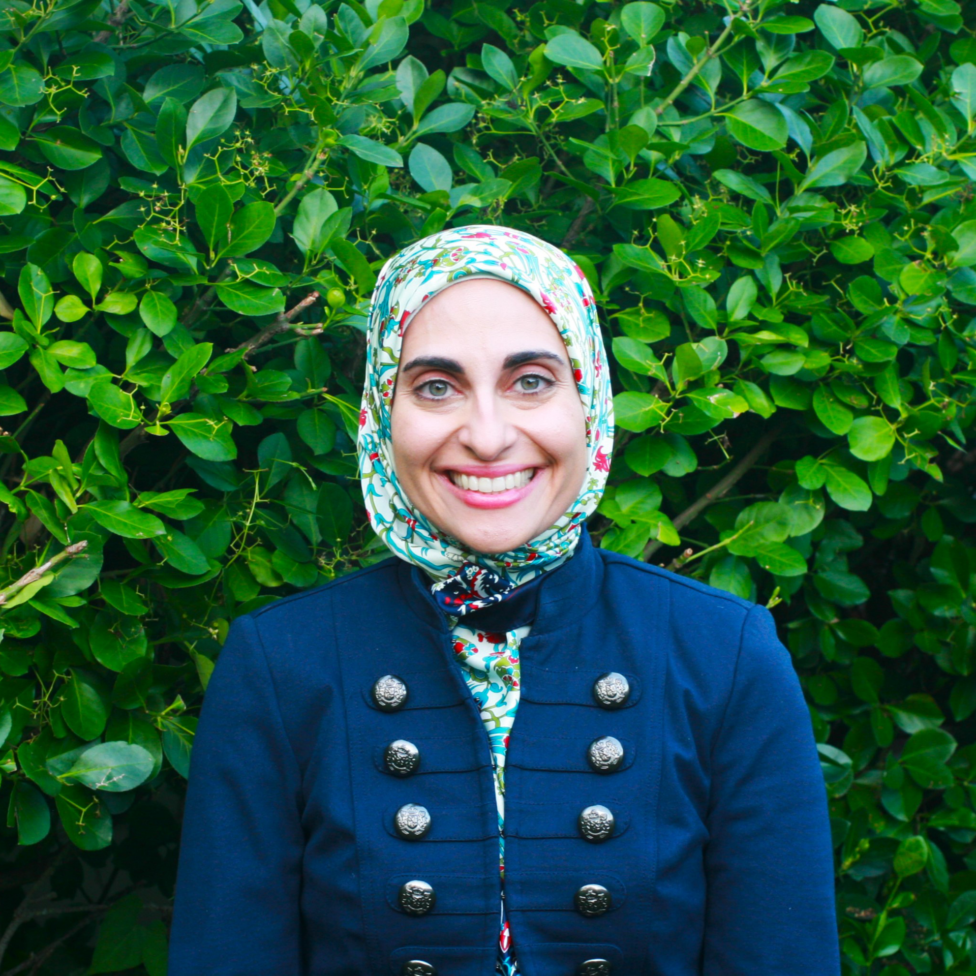 A woman wearing a hijab and a jacket is smiling in front of a brick wall.
