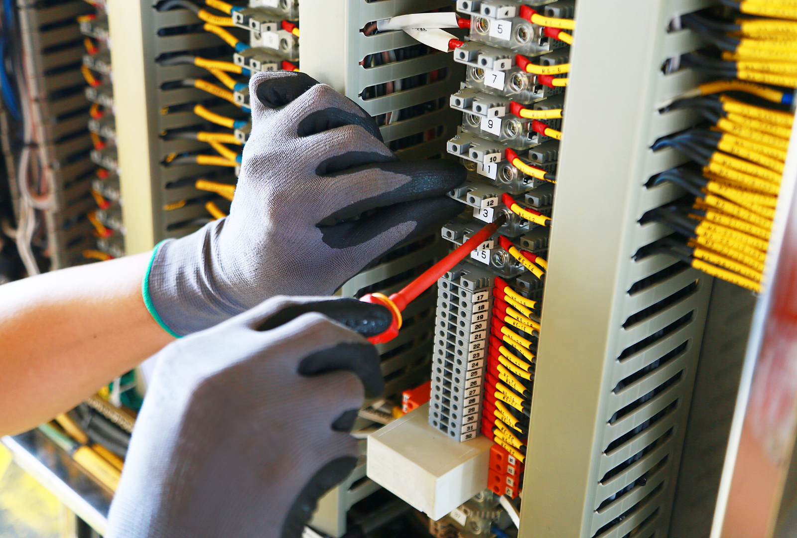 Hands in gloves using a screwdriver to work on a wiring panel with colorful wires.