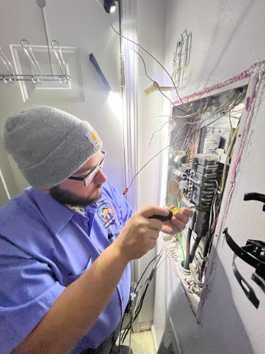 Electrician in a gray beanie and blue shirt working on an open electrical panel.