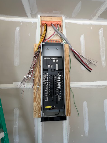 Electrical panel installation: metal box with breakers, wires extending, mounted on wood in a wall.