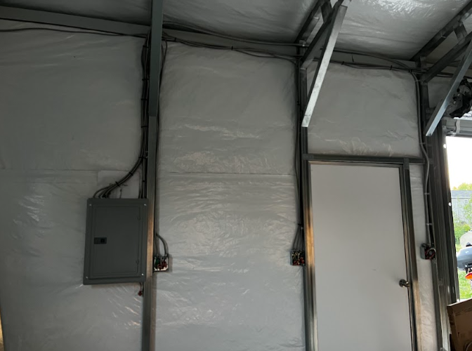 Interior of a metal building with electrical panel, door, and wiring on a gray-walled structure.