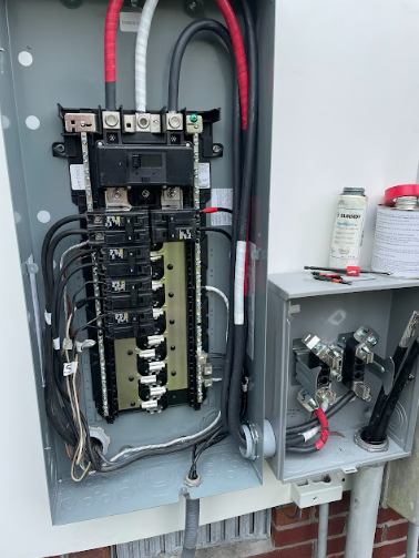 Electrical panel with wires and circuit breakers, mounted on a wall with conduit. Black, red, white wires.