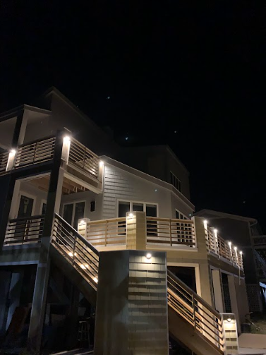Multi-story house exterior at night, lit by warm lights on decks and stairs, against a dark sky.
