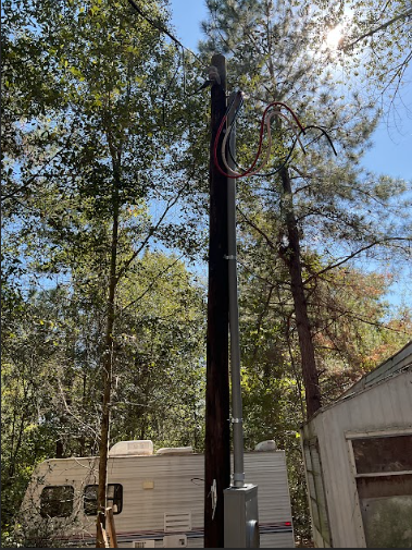 A utility pole with electrical wiring against a backdrop of trees and a trailer.