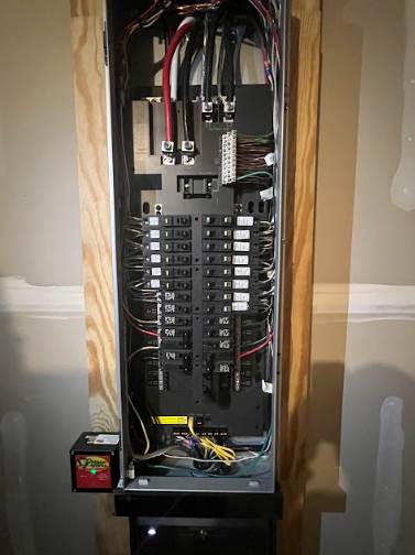 Electrical panel with wires and circuit breakers on a wooden frame, mounted on a wall.