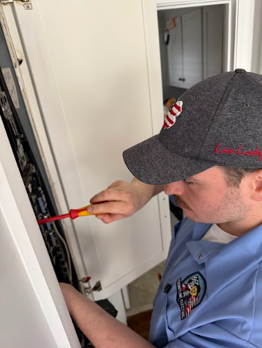 Electrician in a blue uniform and hat working on an electrical panel with a screwdriver.
