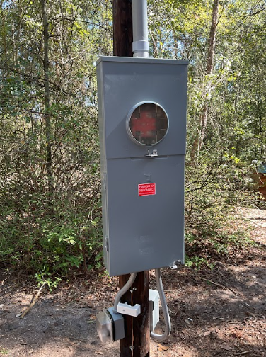 Gray electrical meter box on a wooden pole with conduit, surrounded by trees.