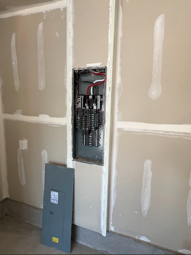 Electrical panel recessed in a drywall wall, panel door on the floor.
