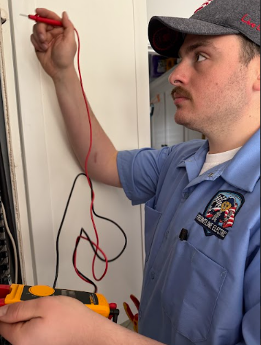 Electrician in blue shirt using a multimeter, testing electrical panel; interior setting.