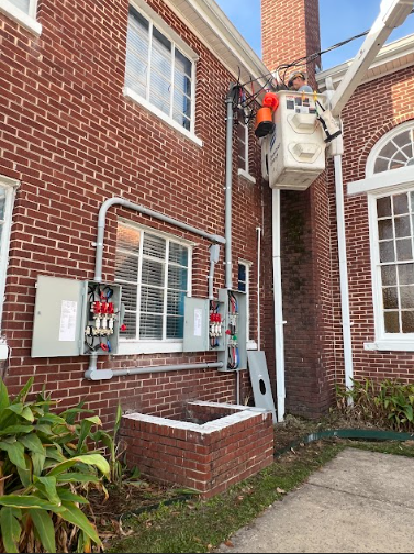 Electrician working on a power transformer mounted on a brick building, near windows and conduit.