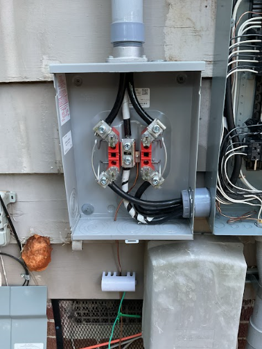 Electrical meter box with wires connected on a house exterior. Gray box, black and white wires, red connectors.