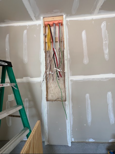 Electrical wires exposed in a wall opening, drywall installation in progress. A green ladder is on the left.