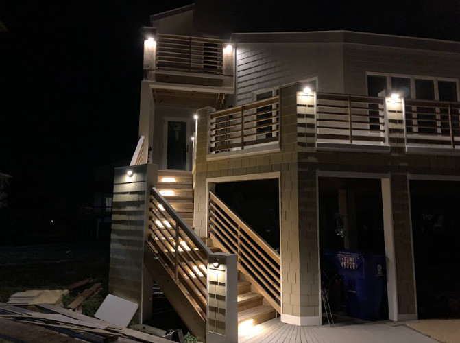 Nighttime exterior view of a house with lit wooden stairs, decks, and siding. Garage door is open.