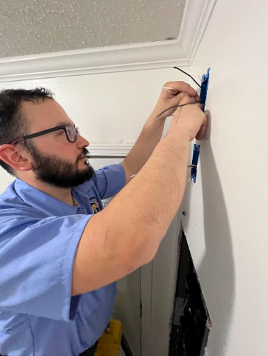 Man in blue shirt using zip ties on wiring in a wall outlet box.