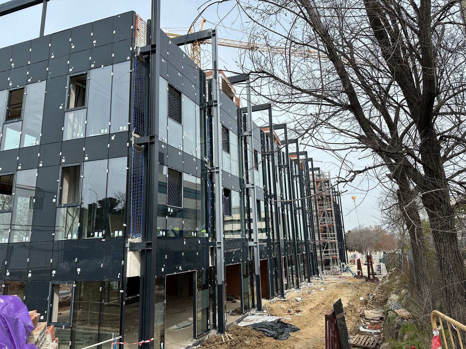 Edificio en construcción con fachada gris oscuro y ventanas de vidrio, camino de tierra y arboles.