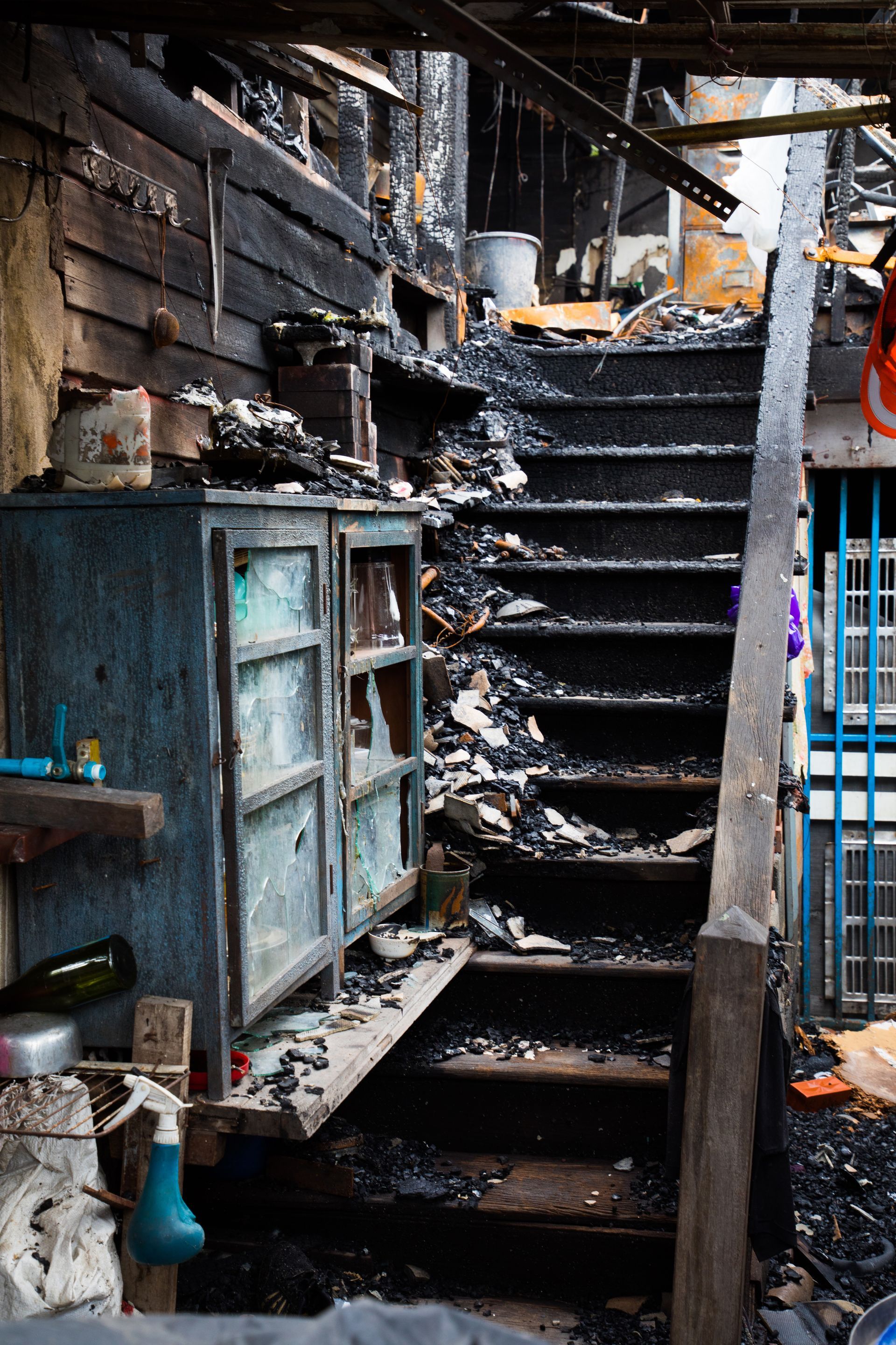 Charred interior: partially destroyed wooden cabinet, stairs, and walls. Debris covers the scene.