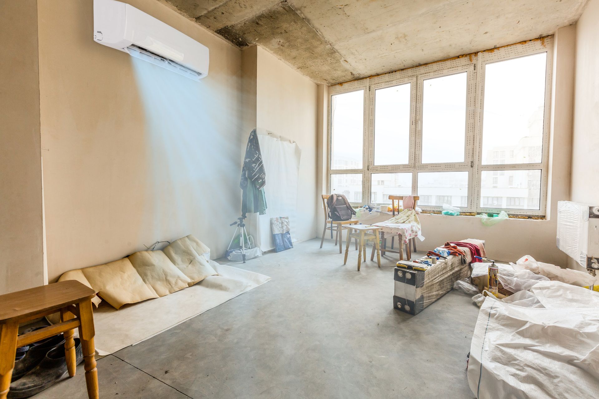 Air conditioner blowing cool air in a messy, unfinished room with a large window.