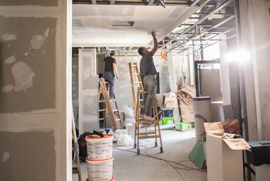 House severely damaged, tilted to the left. Debris and garden on the ground. Driveway on the right. Two construction workers on ladders inside a room under renovation; drywall and tools are visible.