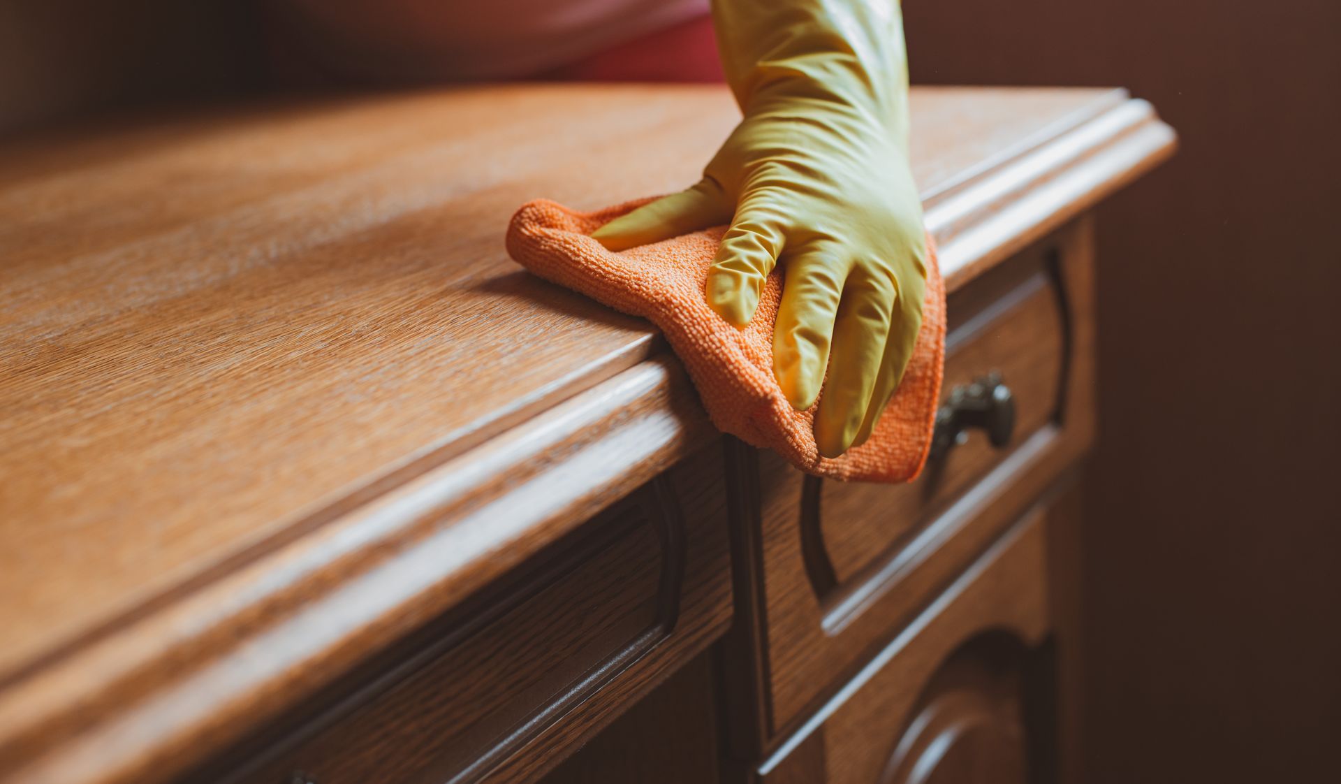 Person in yellow glove wiping wooden furniture with orange cloth. Person in yellow glove wiping wooden furniture with orange cloth.
