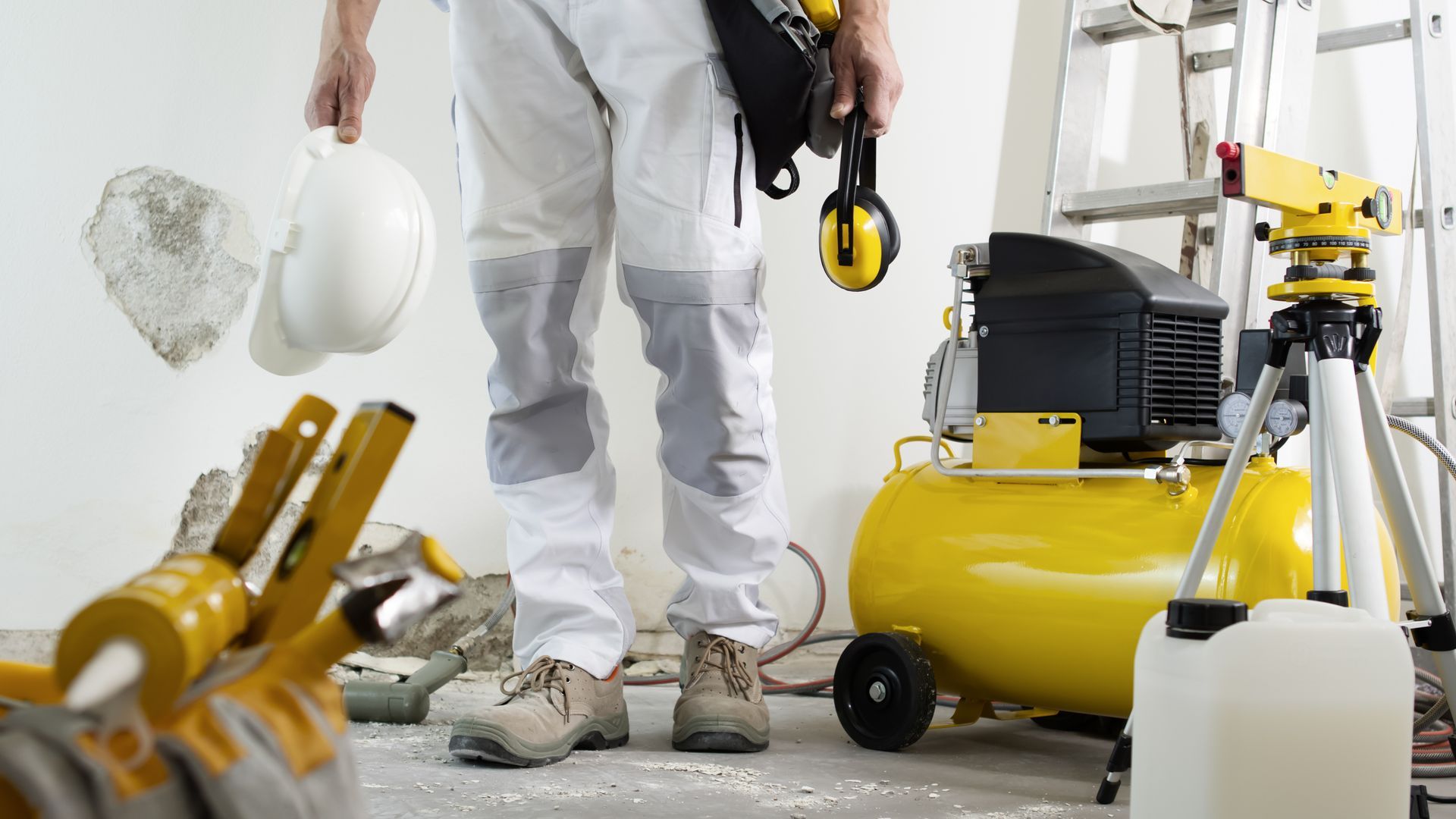 Construction worker in white overalls holding a hard hat, standing near a yellow air compressor and a ladder.