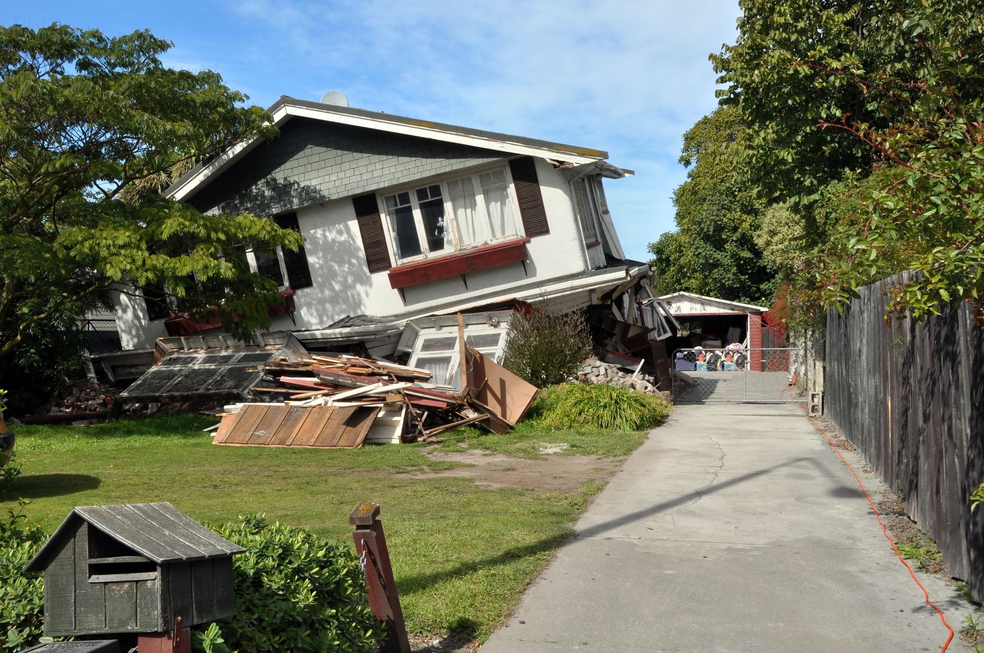 House severely damaged, tilted to the left. Debris and garden on the ground. Driveway on the right. House severely damaged, tilted to the left. Debris and garden on the ground. Driveway on the right.