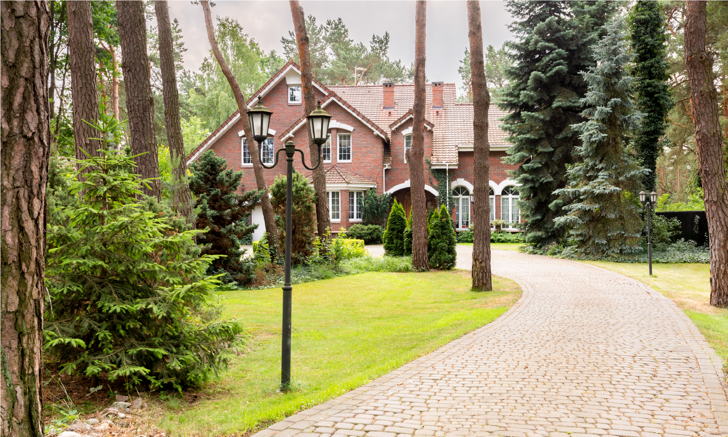 A large brick house is surrounded by trees and a cobblestone driveway.